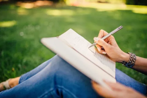 Person sitting on the ground outside on a sunny day writing in a notebook or journal