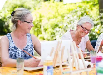 2 ladies with graying hair and glasses sitting in the garden painting on canvas